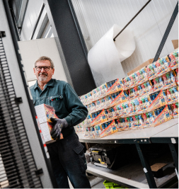 A worker carrying a box in a warehouse with stacks of colorful packaging in the background.